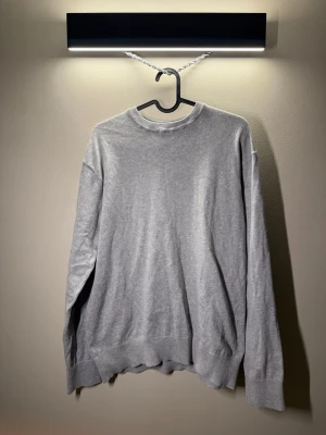 Ljusgrå stickad tröja i bomull - En stilren ljusgrå stickad tröja med rund halsringning och långa ärmar. Tröjan har ribbade muddar vid ärmslut och nederkant, vilket ger en klassisk och avslappnad look. Perfekt att matcha med jeans eller kjol för en enkel och snygg outfit.