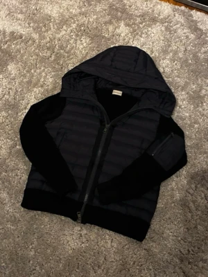 Svart hoodiejacka med quiltad front - Snygg svart hoodiejacka med quiltad front och huva. Jackan har dragkedja framtill, mjuka ärmar och en ficka på ena ärmen. Perfekt för lager på lager och streetwear-stil. Materialet är en mix av vadderad polyester och mjuk bomull.