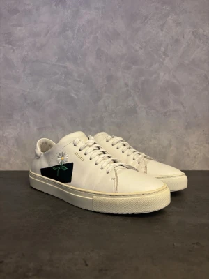 Axel Arigato Clean 90 - Stilrena vita Axel Arigato Clean 90 i läder med broderad blomdetalj och svart panel på sidan. Klassisk låg modell med vit sula och snörning. Perfekta för dig som gillar minimalistisk design med en twist. I bra skick!