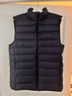 Svart dunväst med hög krage - Snygg svart dunväst med quiltad design och hög krage. Västen har dragkedja framtill och är lätt vadderad för extra värme. Perfekt att slänga över en hoodie eller tröja för en chill och stilren look. Storlek M men sitter som en S