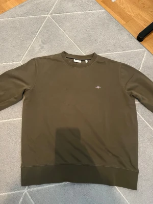Olivgrön sweatshirt från Gant - Säljer en stilren olivgrön sweatshirt från Gant med rund halsringning och diskret logga broderad på bröstet. Tröjan har långa ärmar och ribbade muddar vid ärmslut och nederkant. Perfekt för en avslappnad och trendig look.