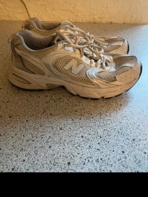 Vita New Balance 530 sneakers - Snygga New Balance 530 sneakers i vitt och silver med meshpaneler och detaljer i syntet. Klassisk N-logga på sidan och bekväm sula. Perfekta för dig som gillar retrostil och sportig vibe.