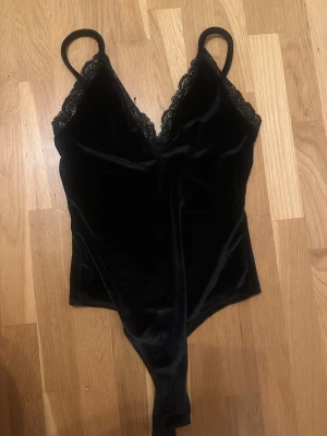 Bodysuit  - Snygg svart body i mjuk sammet med tunna axelband och v-ringning. Kanterna är dekorerade med spets.