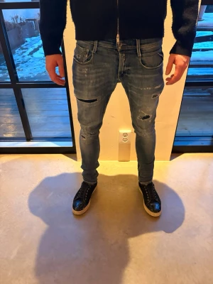 Blå slitna slim jeans replay - Säljer ett par blå slim jeans med slitningar och trasiga detaljer på benen. Jeansen har klassiska fem fickor, dragkedja och knapp framtill samt stora bakfickor. Perfekt för dig som gillar en trendig och avslappnad look. Replay jeans i storlek 29/32 modellnamn anbass