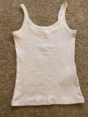 Vit ribbad linnetopp - Säljer en enkel och stilren vit ribbad linnetopp med smala axelband. Toppen är ärmlös och har en tight passform, perfekt att styla till jeans eller kjol. Materialet känns mjukt och stretchigt, vilket gör den superbekväm att bära.