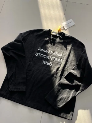 Svart långärmad topp Acne Studios - Cool svart långärmad topp från Acne Studios med vit text 'STOCKHOLM 1996' framtill. Klassisk rund halsringning och relaxed fit. Perfekt till jeans eller kjol för en clean och trendig look.