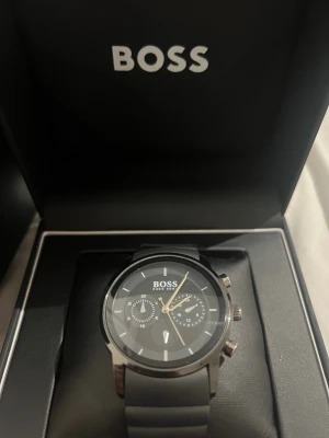 Svart klocka från BOSS med silikonband - Stilren svart klocka från BOSS med rund urtavla och silikonband. Klockan har silverfärgade detaljer, tre subdials och kronograf-funktion. Perfekt accessoar för dig som gillar en modern och clean look.