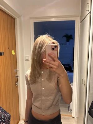 Beige kortärmad kofta med pärlor - Säljer en beige kortärmad kofta med pärlbroderi vid axlarna och vita knappar framtill. Koftan är ribbstickad vid nederkant och ärmslut, och har en croppad passform som är perfekt för att styla med ett par snygga jeans. Använd ett fåtal gånger😇