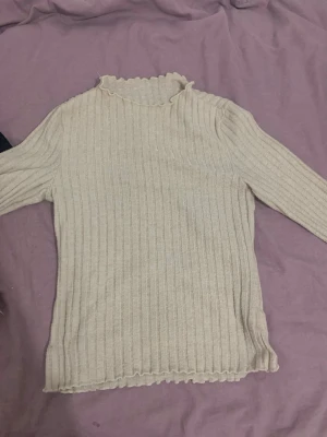 Beige ribbstickad tröja med volangkant - Säljer en beige ribbstickad tröja med långa ärmar och volangkant vid halsringning och nederkant. Tröjan har en smal passform och är perfekt för lager på lager. Materialet känns mjukt och stretchigt.