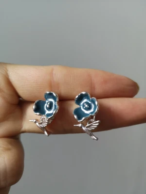 Blå blomörhängen med fågelmotiv - Unika örhängen i form av blå blommor med silverfärgade små fåglar undertill. Blommorna har en glansig finish och örhängena är tillverkade i metall. Perfekta för dig som gillar naturinspirerade accessoarer.