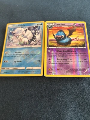 Pokémonkort Alolan Vulpix & Swoobat - Två Pokémonkort: Alolan Vulpix med blå bakgrund och Swoobat med lila och guldfärgad holografisk effekt. Båda korten har tydliga illustrationer och detaljerad design. Perfekt för samlare eller Pokémonfans.
