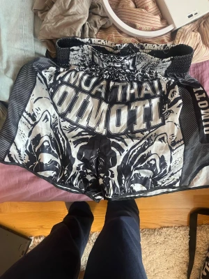 Muay Thai shorts svart/vit tiger - Säljer ett par Muay Thai shorts i svart och vitt med stora tigertryck och texten 'MUAY THAI' samt 'OIMOTI' på framsidan. Shortsen har bred resår i midjan och är tillverkade i glansigt, lätt material som passar perfekt för träning och sparring.