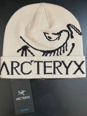Beige mössa från Arc'teryx.! - Snygg beige mössa från Arc'teryx med svart logotyp och text framtill. Mössan är ribbstickad och har uppvikt kant. Perfekt för dig som gillar stilrena accessoarer med outdoor-vibe. Materialet känns mjukt och stretchigt, vilket gör den bekväm att bära.