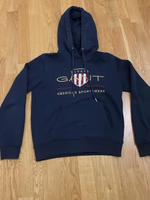 Mörkblå hoodie från Gant - Snygg mörkblå hoodie från Gant med stor logga och amerikansk sköld på bröstet. Klassisk känguruficka framtill, justerbar huva med dragsko och ribbade muddar. Tillverkad i mjukt bomullsmaterial för en bekväm och avslappnad stil. Köpt från Gant butiken i Göteborg 