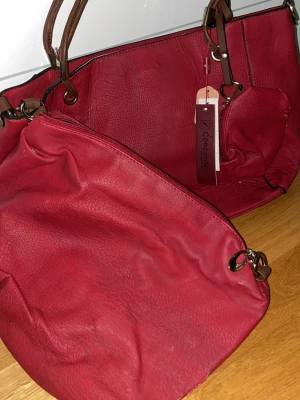 Röd handväska från Conaide - Snygg röd handväska från Conaide i mjukt, strukturerat konstläder. Väskan har bruna handtag, metallringar och en praktisk innerficka. Perfekt för dig som vill ha en rymlig och trendig accessoar med plats för allt du behöver.