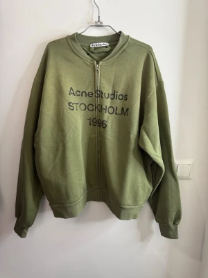 Grön zip hoodie Acne Studios - Snygg grön hoodie med dragkedja från Acne Studios. Tryck på bröstet med texten 'Acne Studios STOCKHOLM 1996'. Oversized passform, ribbade muddar och mjukt material i bomullsblandning. Perfekt för en avslappnad och trendig look.