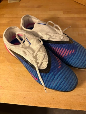 Nike fotbollsskor - Säljer ett par Nike  fotbollsskor i blått, vitt och svart med rosa detaljer. Skorna har en strömlinjeformad design, syntetiskt material och fasta dobbar för bra grepp på planen. Perfekta för dig som vill ha lättviktsskor med snygg look.