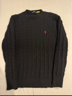 Svart kabelstickad tröja Polo Ralph Lauren - Kabelstickad svart tröja från Polo Ralph Lauren med rund halsringning och den klassiska röda logotypen broderad på bröstet. Tröjan har ribbade muddar vid ärmslut och nederkant, perfekt för dig som gillar stilren och tidlös design. Finns också tags, är helt ny!