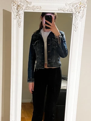 Mörkblå croppad jeansjacka - Säljer en mörkblå croppad jeansjacka med klassiska bröstfickor och knappar framtill. Jackan har en snygg, figurnära passform och är tillverkad i jeansmaterial. Storlek L men skulle säga mer att det är en M. Använd 1 gång.