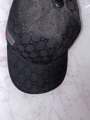 Svart keps från Gucci med monogram - Snygg svart keps från Gucci med det klassiska GG-monogrammet över hela kepsen. På sidan finns en grön och röd rand som ger en ikonisk touch. Kepsen har böjd skärm och knapp på toppen. Perfekt för dig som gillar exklusiva accessoarer.
