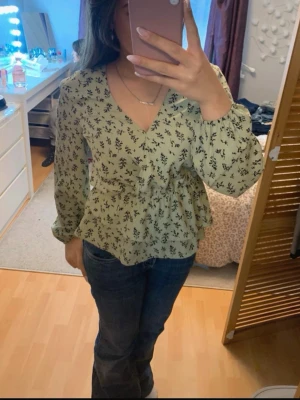 Grön blommig omlottblus - Snygg grön omlottblus med svart blommönster och v-ringning. Blusen har långa ärmar med resår vid handlederna och markerad midja med volang nedtill. Perfekt till jeans eller kjol för en trendig look.