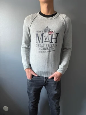 Ljusgrå stickad tröja Morris - Ljusgrå stickad tröja från Morris med tryck på bröstet, inklusive brittisk flagga och texten 'Great Britain'. Tröjan har rund halsringning, ribbade muddar och nederkant. Perfekt för dig som gillar klassisk och stilren design.