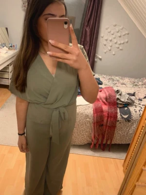 Olivgrön jumpsuit med knytband - Snygg olivgrön jumpsuit med v-ringad omlottfront och korta ärmar. Modellen har ett knytband i midjan och vida ben som ger en relaxed look. Perfekt för dig som gillar stilrena plagg med enkel design.