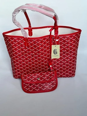 Röd Goyard shoppingväska med mönster - Snygg röd shoppingväska från Goyard med ikoniskt vitt och svart mönster. Väskan har två handtag, öppen topp och kommer med en matchande liten clutch. Tillverkad i tåligt canvasmaterial med läderdetaljer. Perfekt för dig som vill sticka ut med en klassisk designerbag.