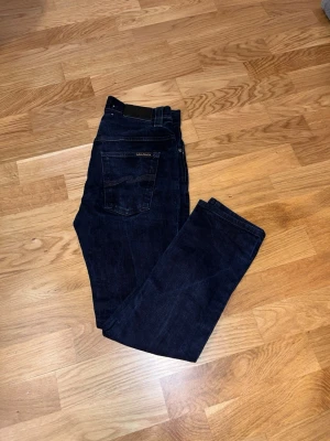 Mörkblå jeans från Nudie Jeans - Säljer ett par mörkblå jeans från Nudie Jeans med klassisk femficksdesign och raka ben. Jeansen har en diskret logga på bakfickan och är gjorda i slitstark denim. Perfekta för en clean och stilren look.