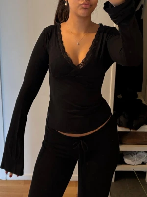Svart långärmad topp med spets - Snygg svart långärmad topp med v-ringning och spetsdetaljer längs urringningen. Modellen är figurnära och har långa ärmar med lätt utsvängd form. Perfekt för dig som gillar en enkel men ändå elegant look.