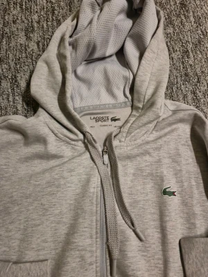 Grå zip-hoodie från Lacoste Sport XXL - Snygg grå zip-hoodie från Lacoste Sport med klassisk krokodillogo på bröstet. Hoodien har huva med dragsko, ribbade muddar och fickor på sidorna. Materialet är mjukt och har meshdetaljer på insidan och sidorna för extra komfort. Storlek xxl