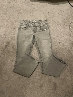 Gråa Jeans - Säljer ett par grå jeans med rak passform och råa, avklippta benslut. Jeansen har klassisk femficksdesign och detaljerade bakfickor med lock. Perfekta för en avslappnad och trendig look. Köpta på Gina Tricot och är i storlek 40.