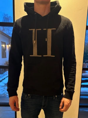 Svart hoodie från les deux - Snygg svart hoodie med stor vit II-logga på bröstet. Klassisk modell med huva och dragsnören samt magficka framtill. Perfekt för en avslappnad och trendig stil. Tillverkad i mjukt bomullsmaterial som känns skönt mot huden.