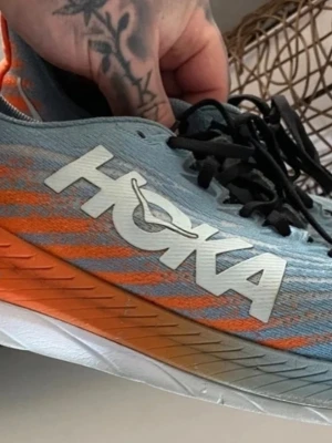 Hoka blå-orange sneakers med snörning - Snygga sneakers från Hoka i ljusblått med orange detaljer och vit sula. Skorna har svart snörning och stor Hoka-logga på sidan. Ovandelen är i syntetmaterial som andas, perfekt för dig som gillar sportig stil. Storlek 