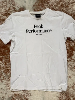 Vit Peak Performance t-shirt - Säljer en vit t-shirt från Peak Performance med klassiskt svart tryck på bröstet. T-shirten har rund hals och korta ärmar, perfekt för en clean och enkel look. Materialet är mjuk bomull som känns skönt mot huden. Snygg att matcha med jeans eller shorts. Toppskick 9,5/10. Bara att höra av sig vid frågor// Carl