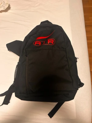 Nike Air svart ryggsäck med röd logga - Snygg svart ryggsäck från Nike med stor röd AIR-logga och swoosh på framsidan. Ryggsäcken har ett stort huvudfack, framficka med dragkedja och sidoficka i mesh. Tillverkad i slitstarkt tyg, perfekt för skolan eller träningen.