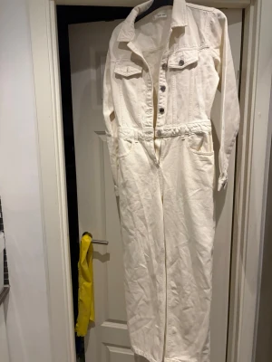 Vit denim jumpsuit med knappar - Säljer en långärmad vit denim jumpsuit med krage och knappar framtill. Den har två bröstfickor med lock, sidofickor och markerad midja. Perfekt för dig som gillar en clean och avslappnad look. Materialet är kraftigt och ger en cool vintage vibe.