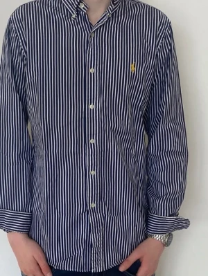 Randig skjorta från Ralph Lauren - Snygg blå och vit randig skjorta från Ralph Lauren med klassisk krage och knappar framtill. Skjortan har lång ärm och ikonisk gul logga broderad på bröstet. Perfekt för en stilren look.