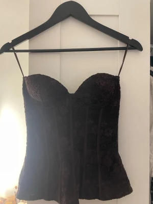 Mörkbrun spetsbody med bygel - Säljer en mörkbrun body. Bodyn är täckt av blommig spets och har markerade sömmar framtill för en snygg siluett. Perfekt för dig som gillar romantiska detaljer och en feminin look.