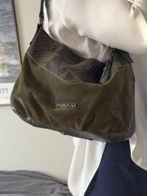 Handväska Ulrika, svensk design - Cool olivgrön handväska från ULRIkA, sevevsk design. Med detaljer i ormmönstrat läder och mocka. Väskan har en bred axelrem och ett rymligt fack med ljus insida och dragkedja. Tillverkad i Italien och perfekt för dig som gillar unika accessoarer.