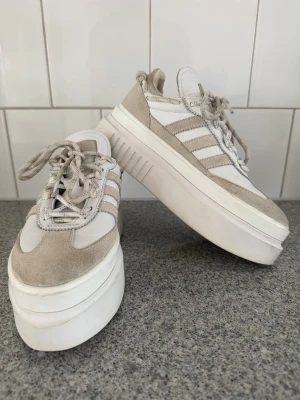 Vita och beige platåsneakers - Ett par vita och beige sneakers från Sdidas (Ivy Park) med kraftig platåsula. Skorna har mockadetaljer, klassiska ränder på sidan och snörning framtill. Perfekta för dig som vill ha extra höjd och en trendig look.