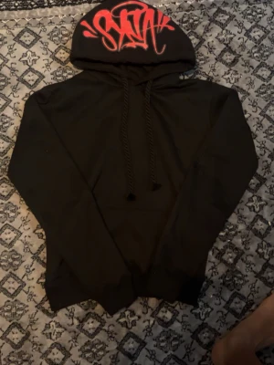 Svart hoodie med röd graffiti-huva - Säljer en svart hoodie med huva och snören. Huvan har en stor röd graffiti-text som ger en streetwear vibe. Hoodien är enkel i modellen och passar perfekt för dig som gillar en avslappnad och cool stil.