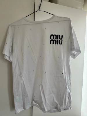 Vit Miu Miu t-shirt med stenar - Säljer en vit t-shirt från Miu Miu med svart broderad logga på bröstet och små glittriga stenar utspridda över hela tröjan. T-shirten har rund hals och korta ärmar. Perfekt för dig som gillar exklusiva detaljer och stilren design.storlek xl men upplevs mindre 