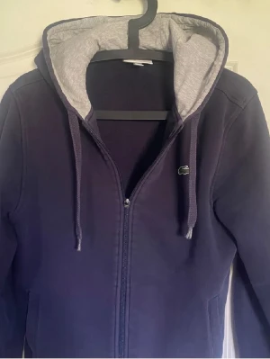 Mörkblå hoodie från Lacoste - Säljer en mörkblå hoodie från Lacoste med dragkedja och grått foder i huvan. Klassisk krokodil-logga på bröstet och justerbara snören i huvan. Tillverkad i mjukt bomullsmaterial som är skönt att ha på sig. Perfekt för dig som gillar stilrena och sportiga plagg.