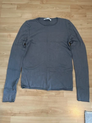 Grå långärmad tröja från Zara - En stilren grå långärmad tröja från Zara med rund halsringning. Tröjan är tillverkad i ett mjukt och tunt material som passar perfekt för lager-på-lager. Enkel design utan mönster eller tryck, vilket gör den lätt att matcha med olika outfits.