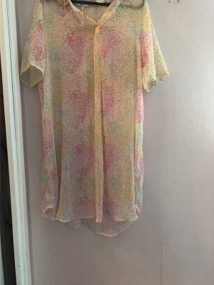 Mönstrad blus från Ellos - Luftig blus från Ellos i transparent material med korta ärmar. Blusen har ett färgglatt mönster i pastellfärger som rosa, gul och mintgrön. V-ringad hals och lös passform gör den perfekt för en avslappnad stil.