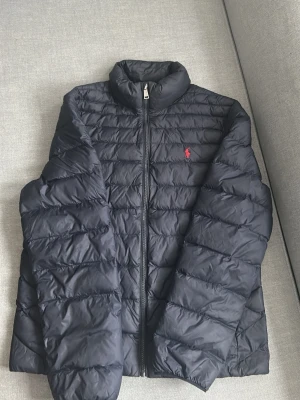 Marinblå Lightweight jacka från Ralph Lauren - Snygg marinblå Lightweight från Ralph Lauren med diskret röd logga på bröstet. Jackan har hög krage, dragkedja framtill och klassisk design. Perfekt för en tunnare jacka till våren och ger en clean look. Materialet är lätt och isolerande. En söm saknas på ena armen och kan behöva sys därav priset.
