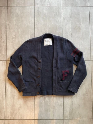 Mörkblå cardigan från Abercrombie & Fitch  - Snygg mörkblå cardigan från Abercrombie & Fitch med knappar framtill, två fickor och en stor röd F-applikation på ena sidan. Ärmen har två röda ränder och koftan är stickad i ett tjockare material. Perfekt för dig som gillar collegeinspirerad stil. Passar strl: S-M