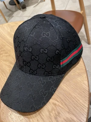 Svart Gucci keps med GG-mönster - Säljer en svart keps från Gucci med det klassiska GG-monogrammet över hela kepsen. På sidan finns en grön och röd rand som ger den en ikonisk touch. Kepsen har böjd skärm och är tillverkad i ett slitstarkt tygmaterial.