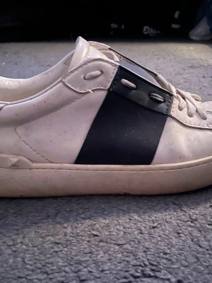 Vit och blå sneaker från Valentino - Snygg vit sneaker från Valentino med en bred svart diagonal detalj på sidan. Skon har låga kanter, vita snören och metalliska öljetter. Tillverkad i läder med klassisk siluett och diskret logga på plösen.